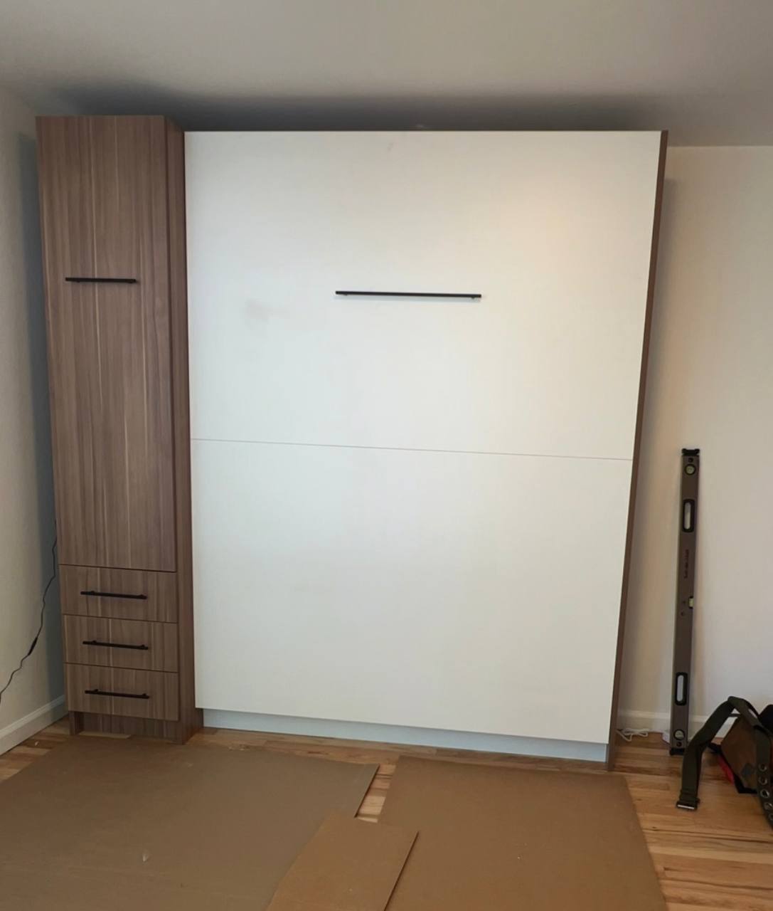 A wall-mounted Murphy bed or fold-down bed panel in white with wood trim, shown installed in a room with wood flooring, cardboard protection sheets, and a level standing on the floor.